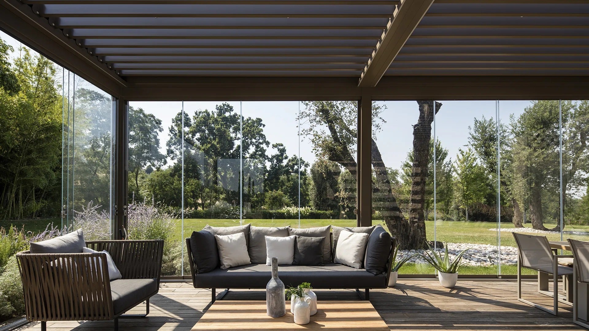 Vetrate panoramiche antivento scorrevoli installate in pergola con vista sul giardino, zona living esterna arredata, proposta Domotea Milano.