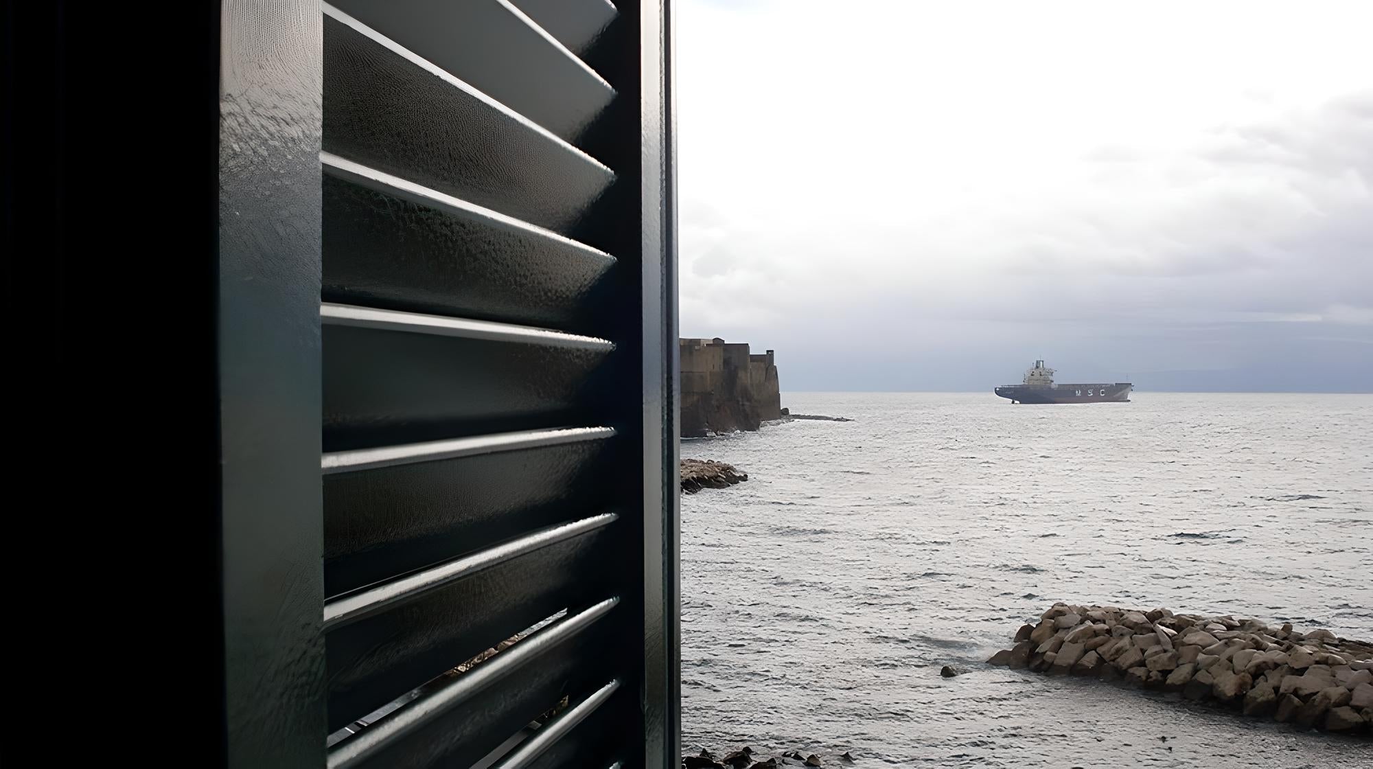 Dettaglio di una persiana in alluminio verniciato con lamelle orientabili, affacciata sul mare con vista su scogliera e nave cargo all’orizzonte.