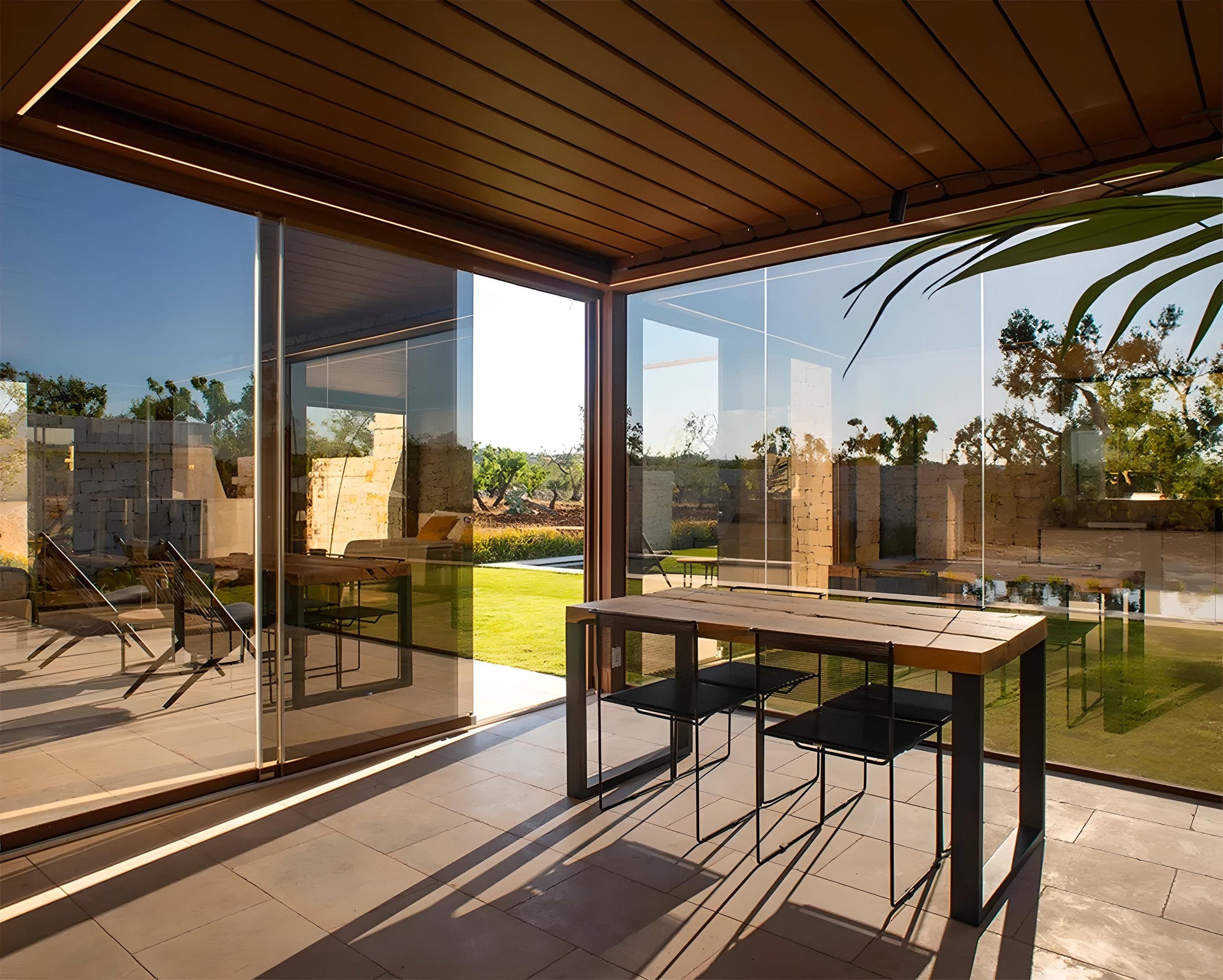 Interno di una pergola bioclimatica con chiusure in vetro e vista su giardino, esempio di spazio esterno valorizzato con soluzioni su misura