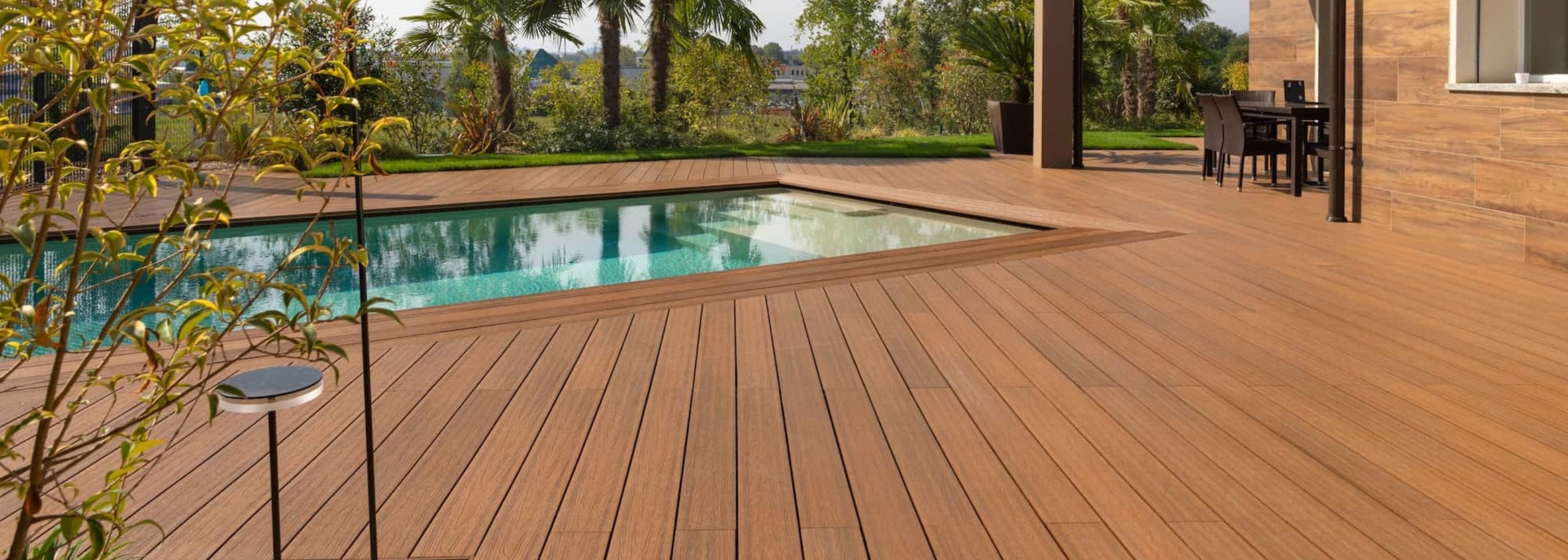 Decking per esterni effetto legno installato attorno a una piscina residenziale, in un giardino con palme e zona pranzo.