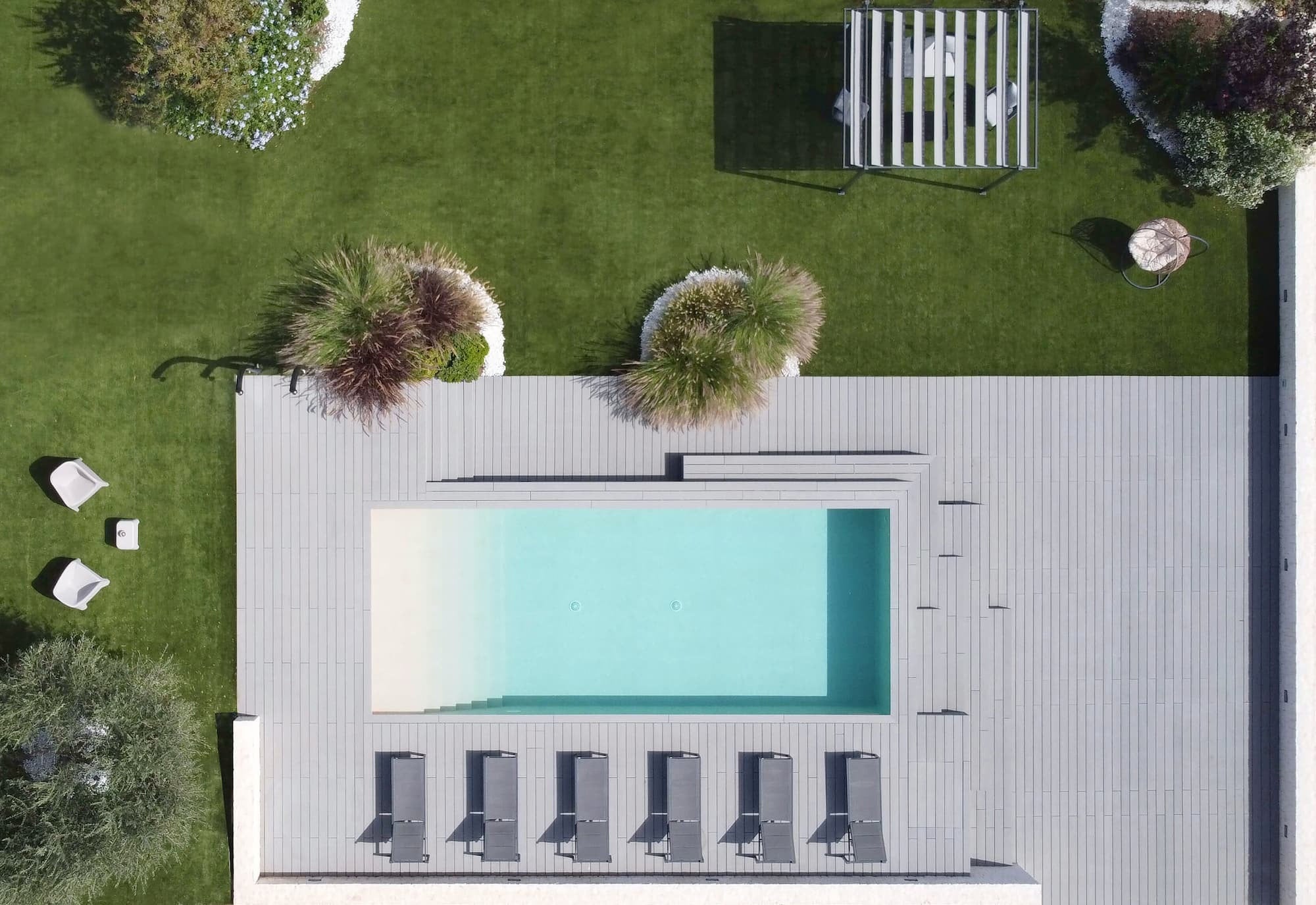 Vista aerea di una piscina con pavimentazione per esterni (decking) effetto legno grigio, sei lettini prendisole allineati e prato verde circostante con elementi d’arredo e vegetazione ornamentale.