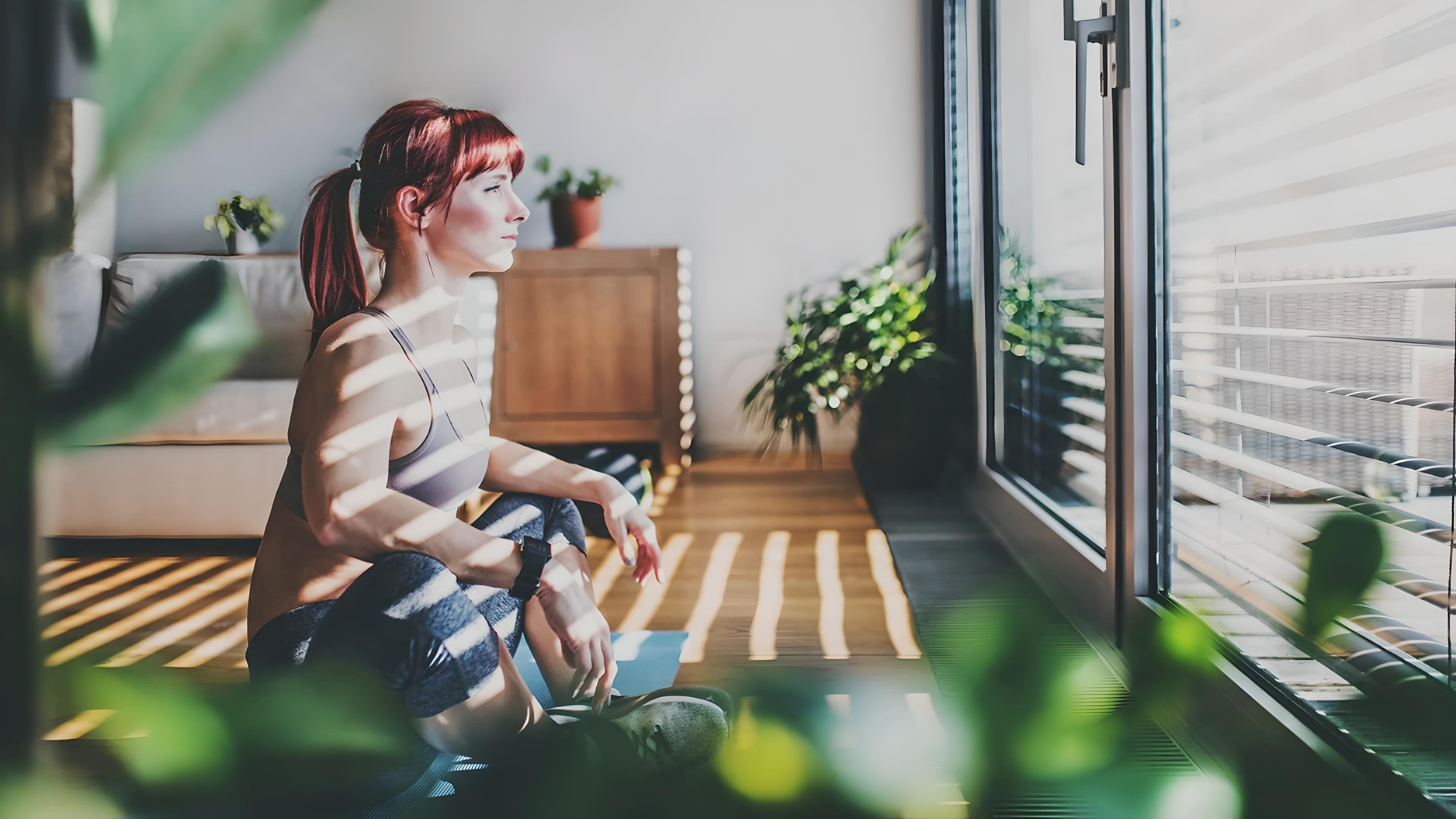 Donna seduta su tappetino da yoga in una stanza luminosa con tende a lamelle orientabili e luce filtrata