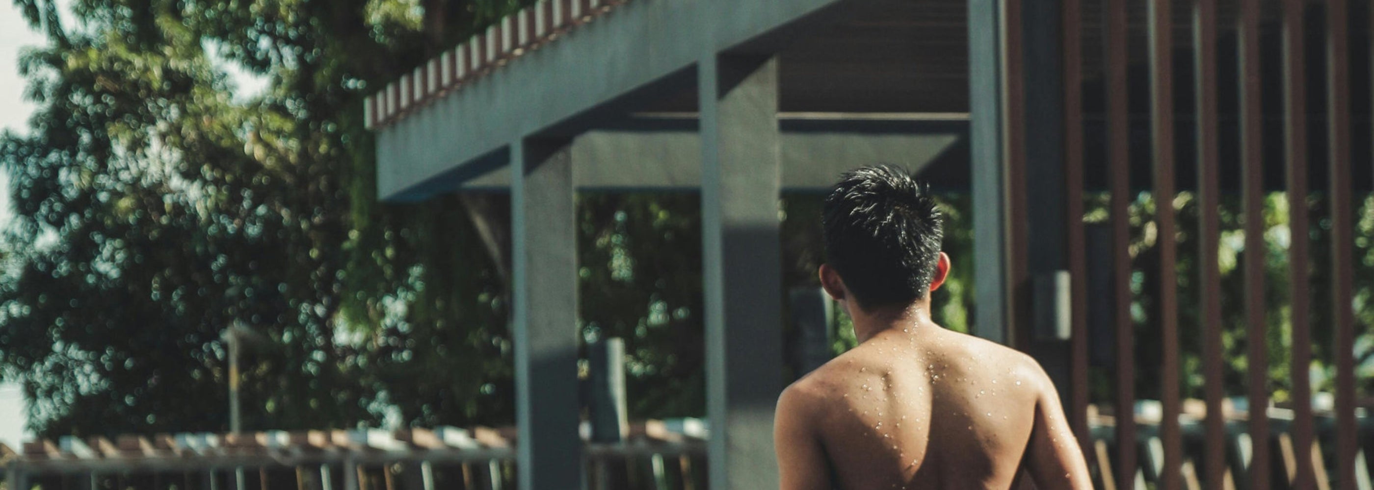 Vista laterale di una piscina con bordo a sfioro e pergola in alluminio, con una persona che cammina lungo il bordo dell’acqua in un contesto residenziale immerso nel verde.