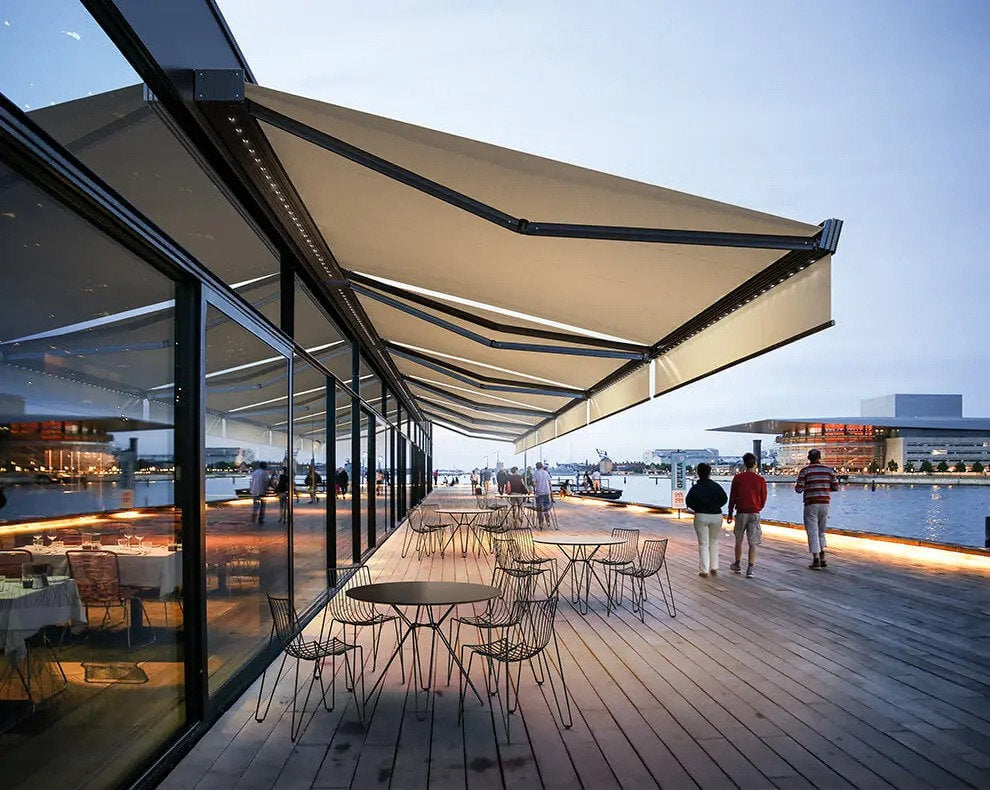 Tende da sole a bracci installate lungo la vetrata di un ristorante affacciato sul porto, con sedute all’aperto e passaggio pedonale illuminato al tramonto.