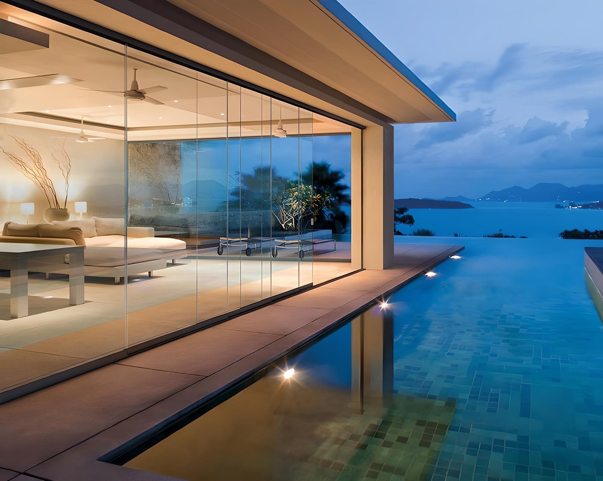 Ambiente serale con piscina e vetrate panoramiche scorrevoli che separano il soggiorno da un terrazzo vista mare, integrate in una villa contemporanea affacciata su un paesaggio costiero.