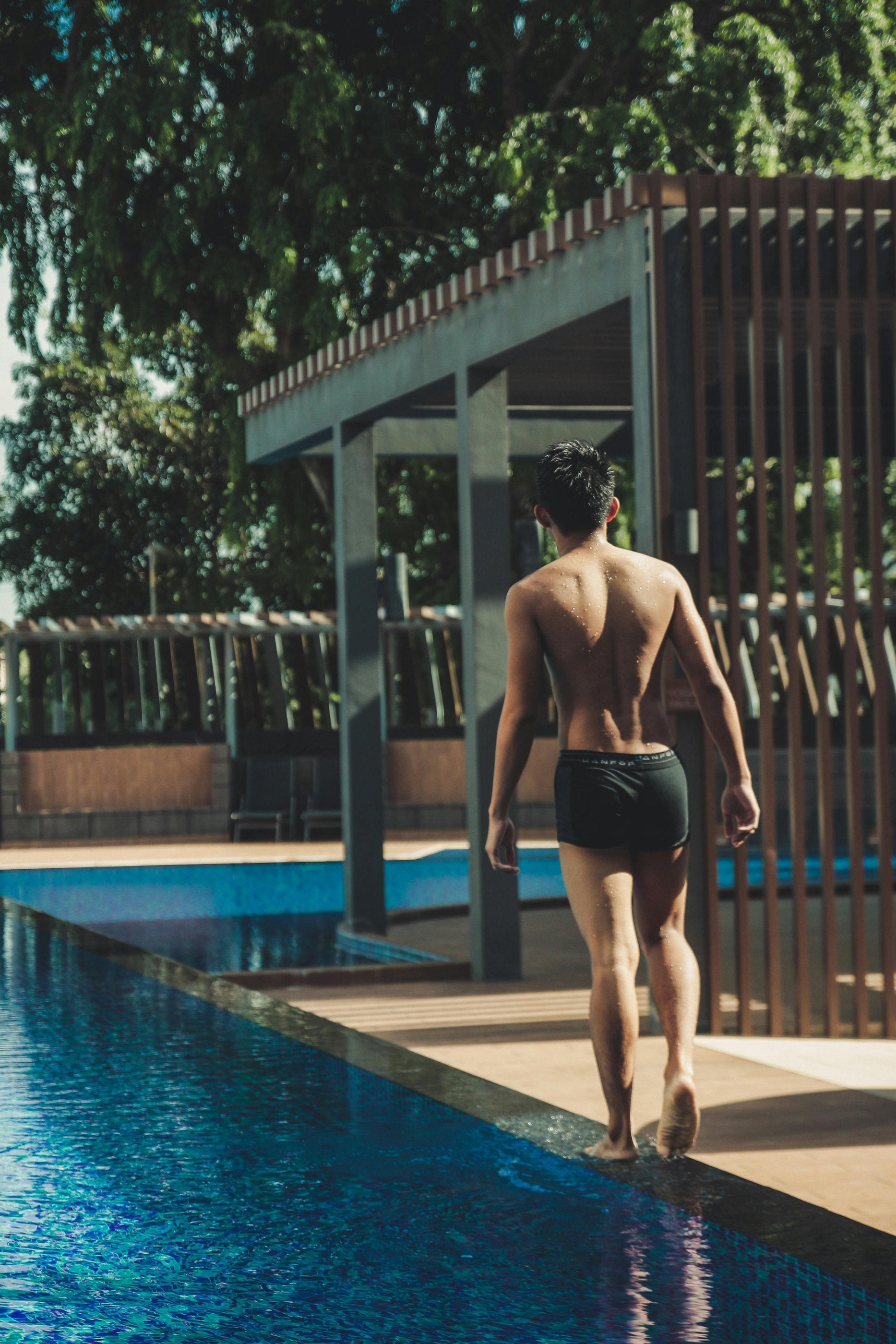 Vista laterale di una piscina con bordo a sfioro e pergola in alluminio, con una persona che cammina lungo il bordo dell’acqua in un contesto residenziale immerso nel verde.