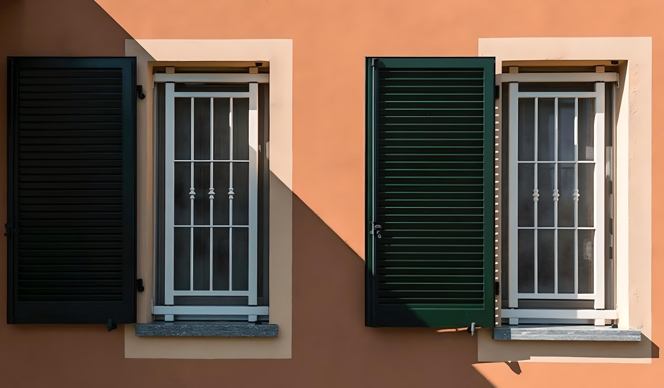 Inferriate di sicurezza bianche installate su finestre con persiane verdi in un edificio dal colore arancione, riprese in luce solare diretta.