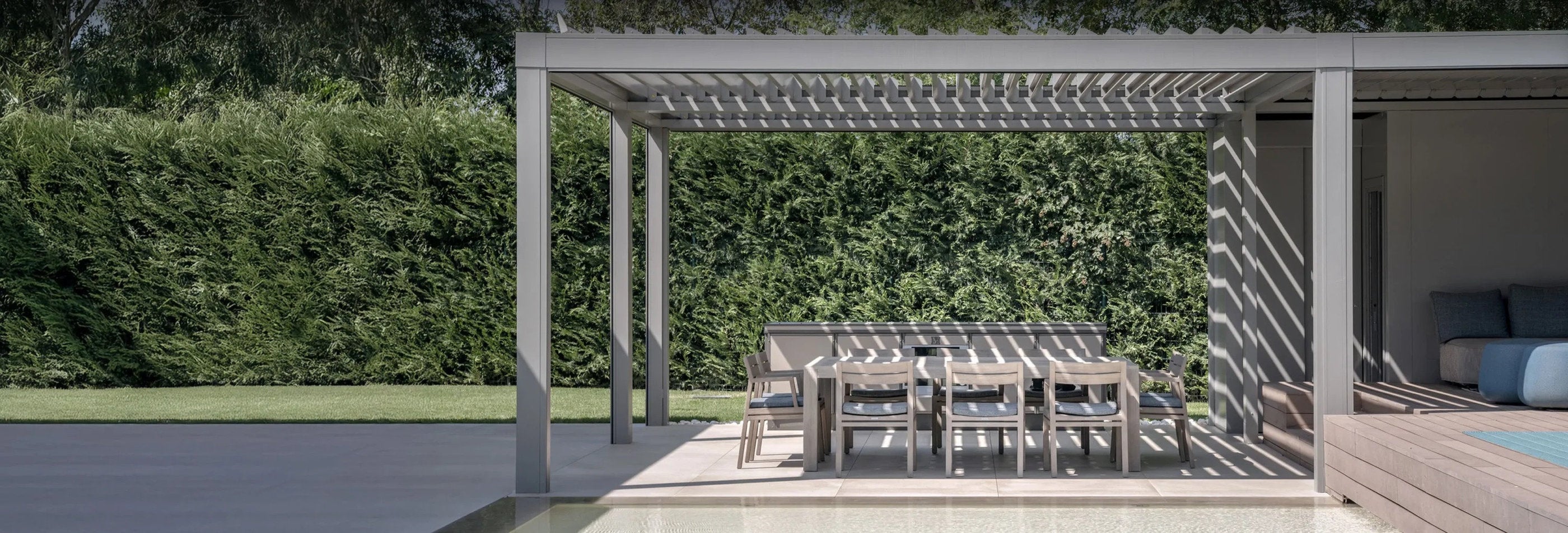 Pergola con lamelle orientabili installata a bordo piscina, ombreggia una zona pranzo all'aperto immersa nel verde.